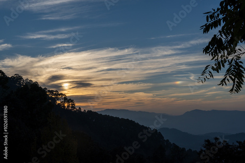 Textured clouds in orange and white hues like brush strokes on a canvas.Blue skies with textured clouds.Silhouette of layered mountains and trees creates a perfect sunset landscape in mountain village