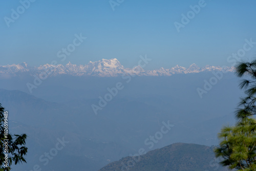 Hazy mountain landscape with faint layers of green mountains.Snow covered peaks of great Himalaya range is clearly visible in the distance.Captured through partially visible pine trees on either side. photo