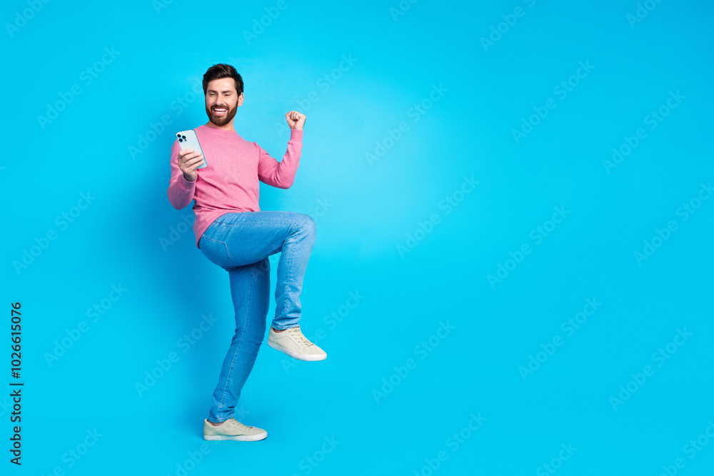Joyful man celebrating success in fashion with casual wear while holding smartphone against vibrant blue background Joyful man celebrating success in fashion with casual wear while holding smartphone against vibrant blue background