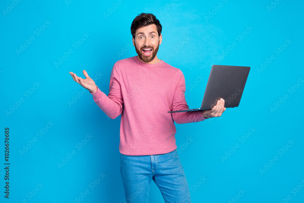 Surprised man holding laptop in pink sweater and jeans against blue background, showcasing excitement and technology