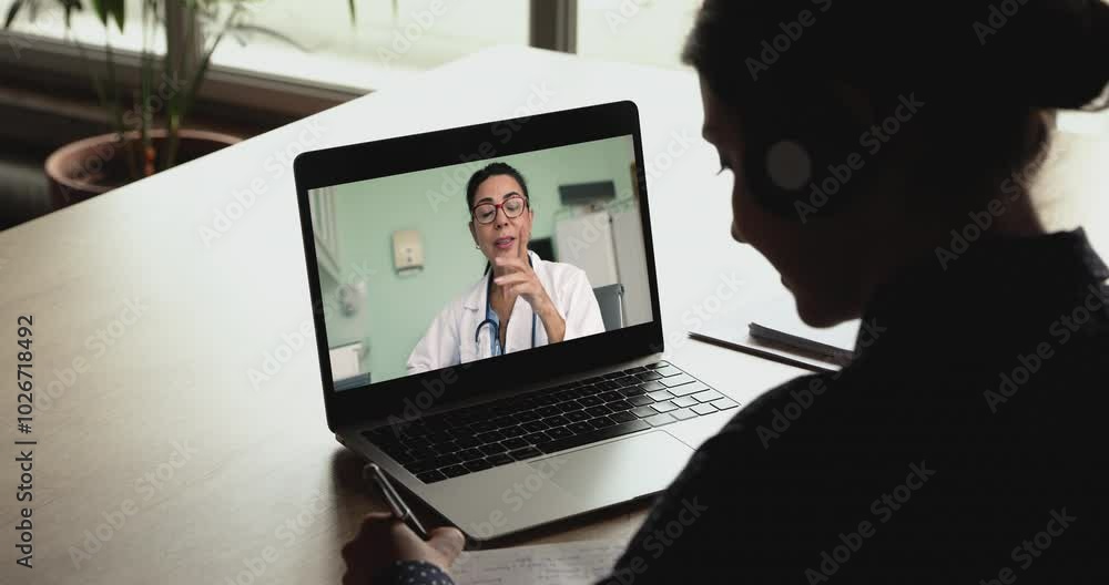Aged woman therapist consulting client by videocall, laptop screen view over female patient shoulder, sit at desk, writing health recommendation. Virtual communication, global connectivity, telehealth