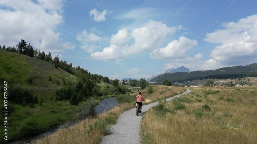 Woman on bicycle enjoys scenic ride on sunny day by river with mountains in distance. Crowsnest Pass, Alberta, Canada. Woman on bicycle enjoys scenic ride on sunny day by river with mountains in distance. Crowsnest Pass, Alberta, Canada.