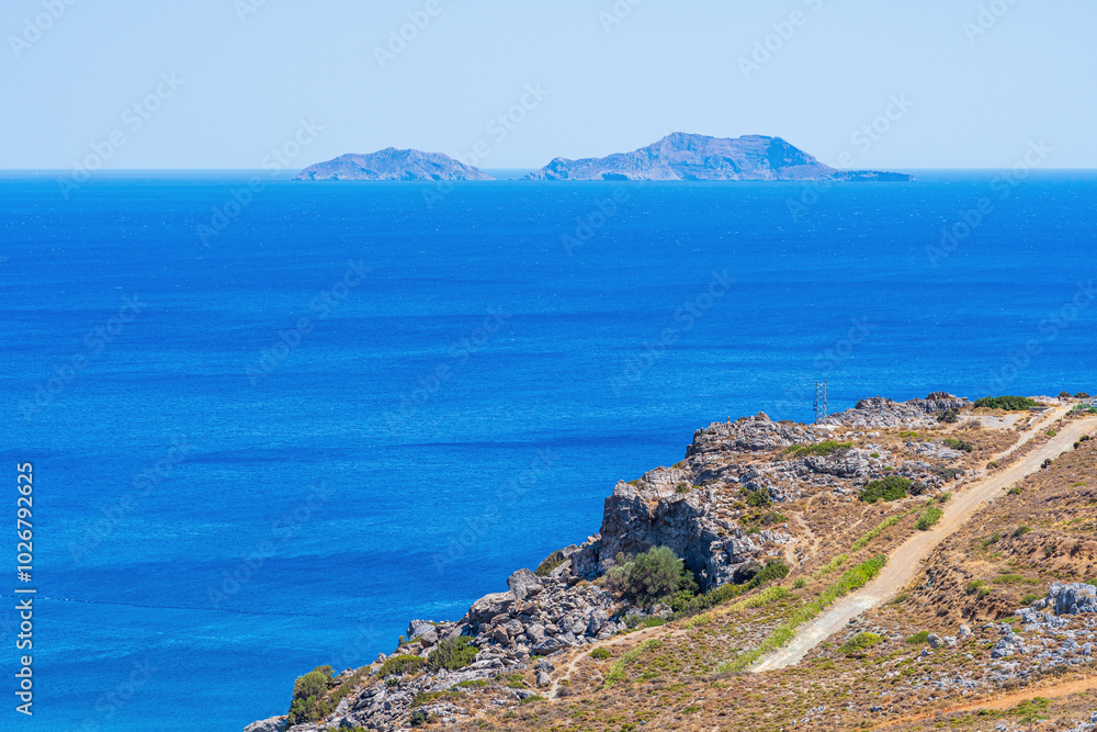 Barren rocky mountainside on a beach with calm sea, two islands in the distance, sunshine, telephoto Barren rocky mountainside on a beach with calm sea, two islands in the distance, sunshine, telephoto