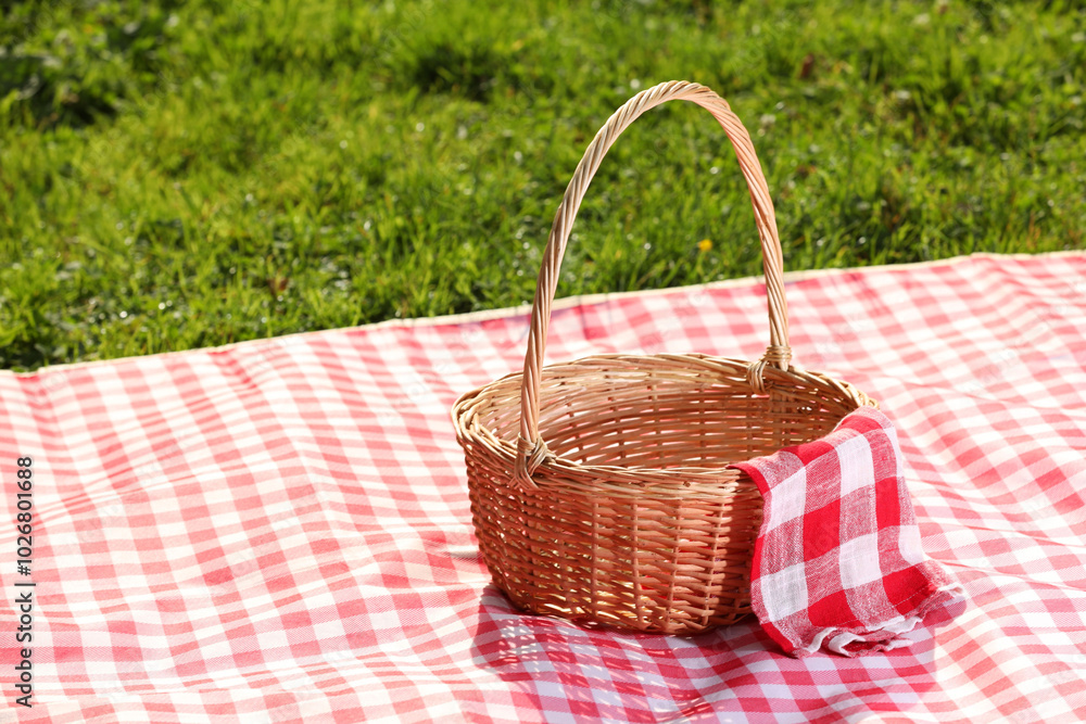 One picnic wicker basket with checkered napkin and blanket on green grass. Space for text One picnic wicker basket with checkered napkin and blanket on green grass. Space for text