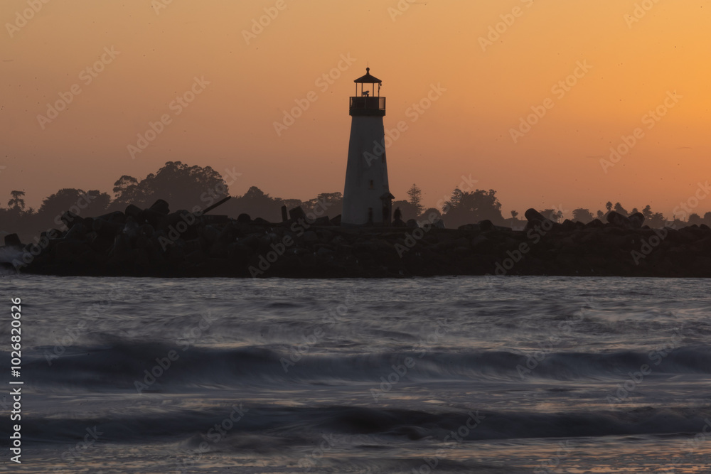 A classic lighthouse stands tall against a vibrant sunset. The silhouette of the lighthouse is stark against the fiery orange and yellow sky. Waves crash against the rocky shore in the foreground A classic lighthouse stands tall against a vibrant sunset. The silhouette of the lighthouse is stark against the fiery orange and yellow sky. Waves crash against the rocky shore in the foreground