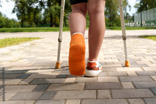 person using crutches walks on a pathway with a bright orange cast, surrounded by greenery