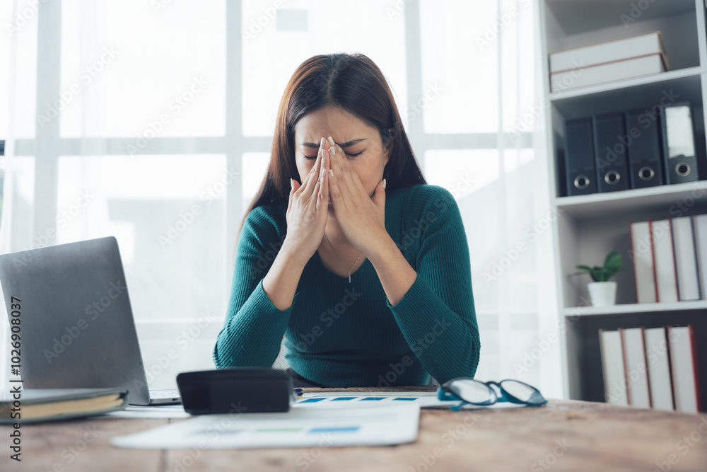 Overwhelmed Businesswoman: Stressed and frustrated, a young businesswoman sits at her desk, hands covering her face, amidst paperwork and a laptop. Overwhelmed Businesswoman: Stressed and frustrated, a young businesswoman sits at her desk, hands covering her face, amidst paperwork and a laptop.