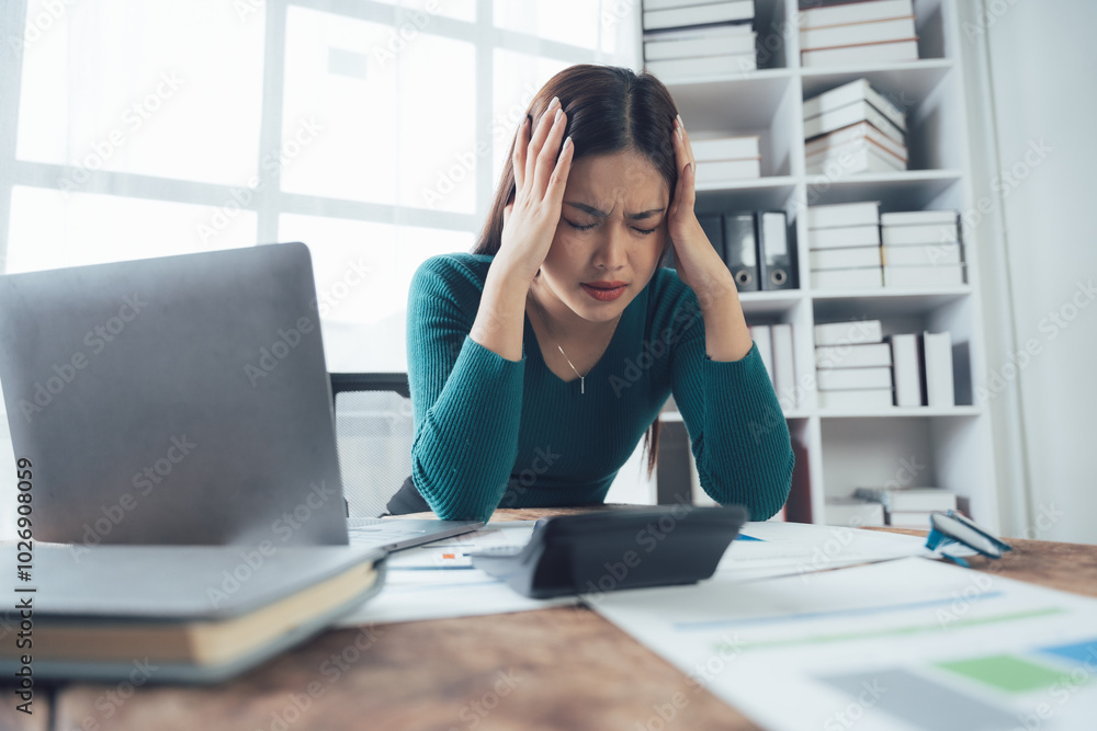 Overwhelmed Businesswoman: Stressed and frustrated, a young businesswoman sits at her desk, hands to her head, surrounded by paperwork and a calculator. Overwhelmed Businesswoman: Stressed and frustrated, a young businesswoman sits at her desk, hands to her head, surrounded by paperwork and a calculator.