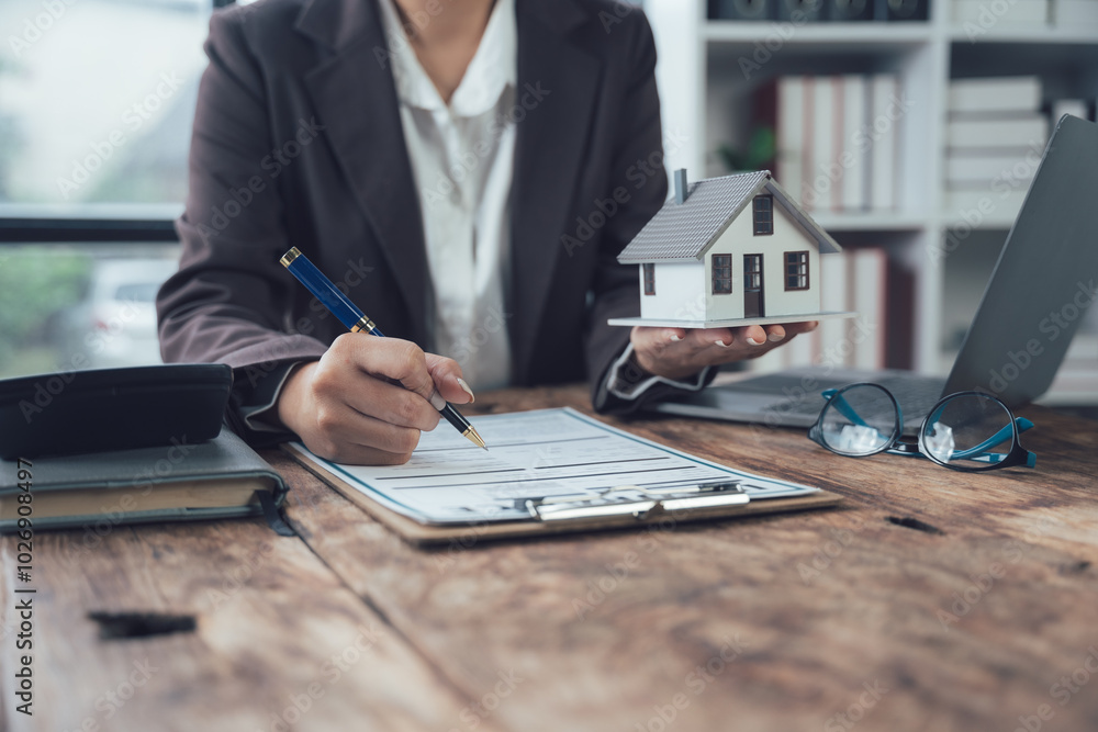 Fototapeta premium Real Estate Agent Signing Contract: A close-up shot of a real estate agent's hand signing a contract while holding a small model home, symbolizing a successful transaction and new beginnings.