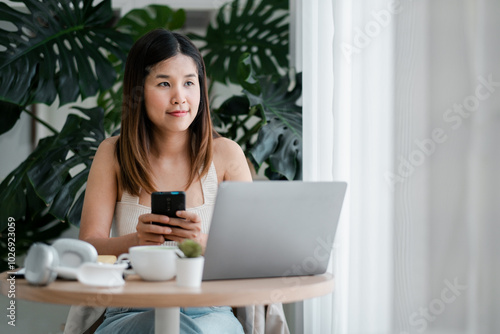 A young woman using a laptop and smartphone while working remotely in a modern home office with natural light and green plants. Free Download A young woman using a laptop and smartphone while working remotely in a modern home office with natural light and green plants. - GFXLoad
