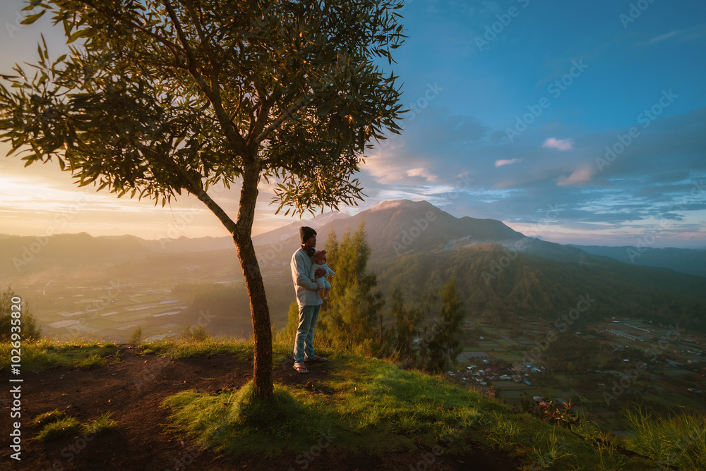 A man and a young child are standing together under the shade of a large tree on top of a hill overlooking the scenery around them A man and a young child are standing together under the shade of a large tree on top of a hill overlooking the scenery around them
