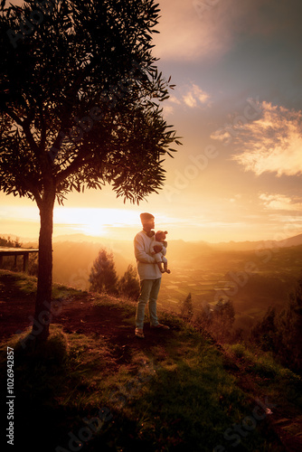 A man is gently holding a dog in front of a large tree as the sun sets, painting the sky with beautiful hues of orange and pink