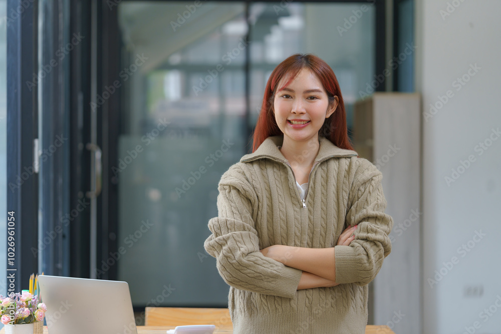 Confident young businesswoman keeping her arms crossed and looking at the camera while standing at her workspace. Confident young businesswoman keeping her arms crossed and looking at the camera while standing at her workspace.