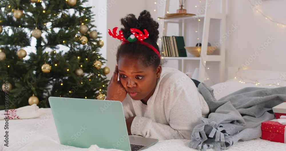 Young smiling african american girl lying on bed in modern bedroom with decorated Christmas fir tree and chatting with a friend using laptop having online video call at home. 4k video. Stock 비디오 | Adobe Stock