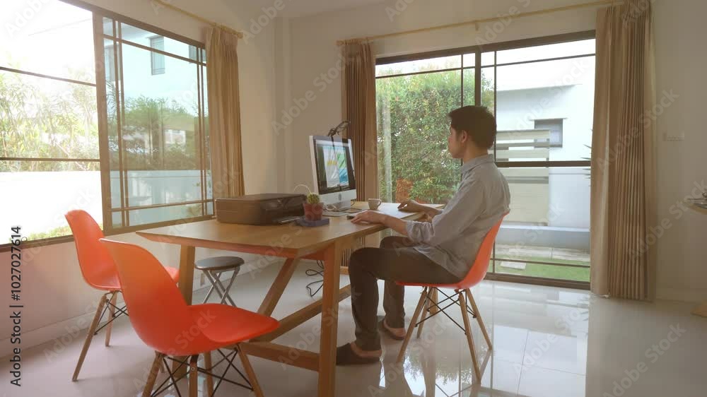 A man is sitting at a desk in a room with a computer monitor and a printer. He is wearing a blue shirt and is looking at the computer screen. The room has a modern and professional feel to it A man is sitting at a desk in a room with a computer monitor and a printer. He is wearing a blue shirt and is looking at the computer screen. The room has a modern and professional feel to it