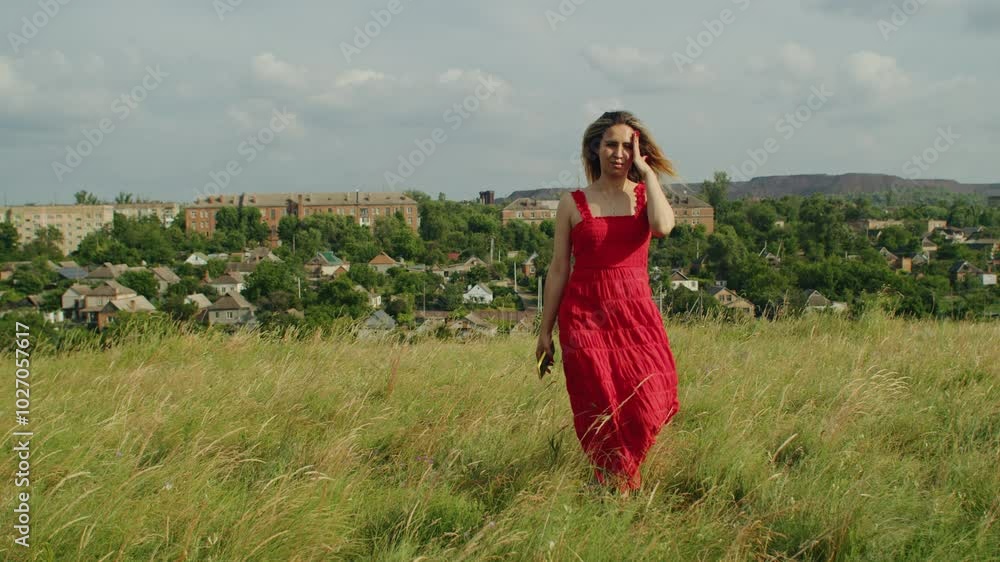 Woman in a red dress walking through a grassy field with a rural town ...