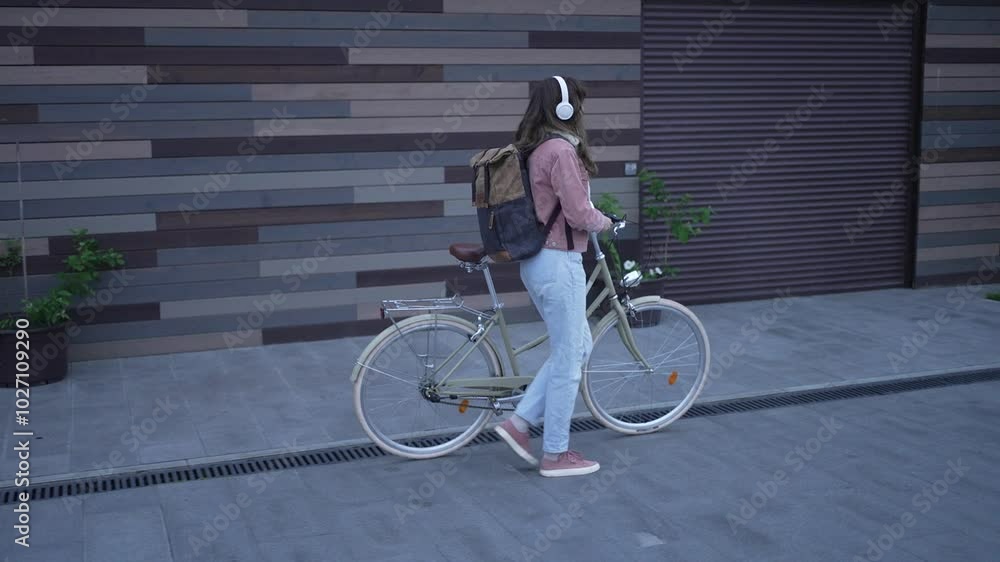 Woman strolling with a bicycle on a contemporary urban street while listening to music through headphones. Relishing the fresh air in a chic neighborhood Woman strolling with a bicycle on a contemporary urban street while listening to music through headphones. Relishing the fresh air in a chic neighborhood