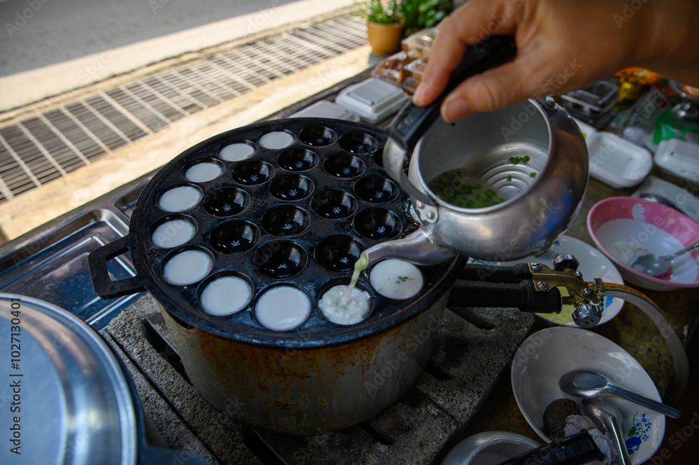 A vendor is using a kettle with khanom krok ingredients to pour into a hot, pitted pan until cooked into a Thai dessert called khanom krok.