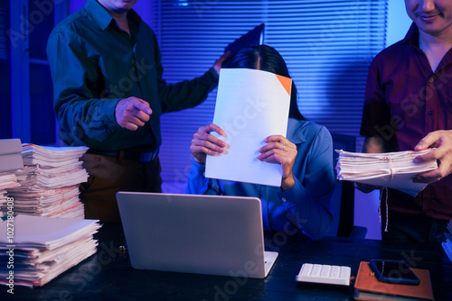 Two men bring a large workload to a woman at her desk in the office. She feels stressed and pressured as she checks documents, working overtime at night and feeling tired photo