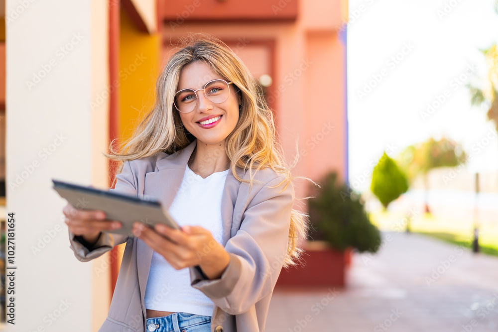 Pretty blonde Uruguayan woman holding a tablet at outdoors with happy expression Pretty blonde Uruguayan woman holding a tablet at outdoors with happy expression