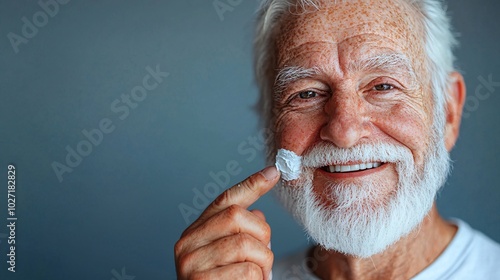 Western elderly man applying eye cream, smiling confidently at the camera, isolated on a neutral gray background Stock Photo with side copy space