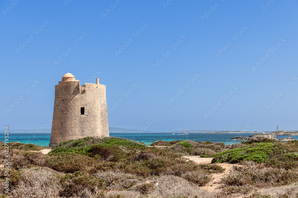 Ibiza ancient coastal tower Torre de ses Portes stands against a backdrop of azure waters and clear skies. Lush greenery and distant sailing vessels add depth to the tranquil and picturesque landscape Ibiza ancient coastal tower Torre de ses Portes stands against a backdrop of azure waters and clear skies. Lush greenery and distant sailing vessels add depth to the tranquil and picturesque landscape