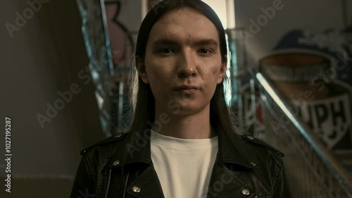 Young, handsome guy in a black rocker jacket and long hair stands in the entrance of a house in a disadvantaged neighborhood and looks at the camera.