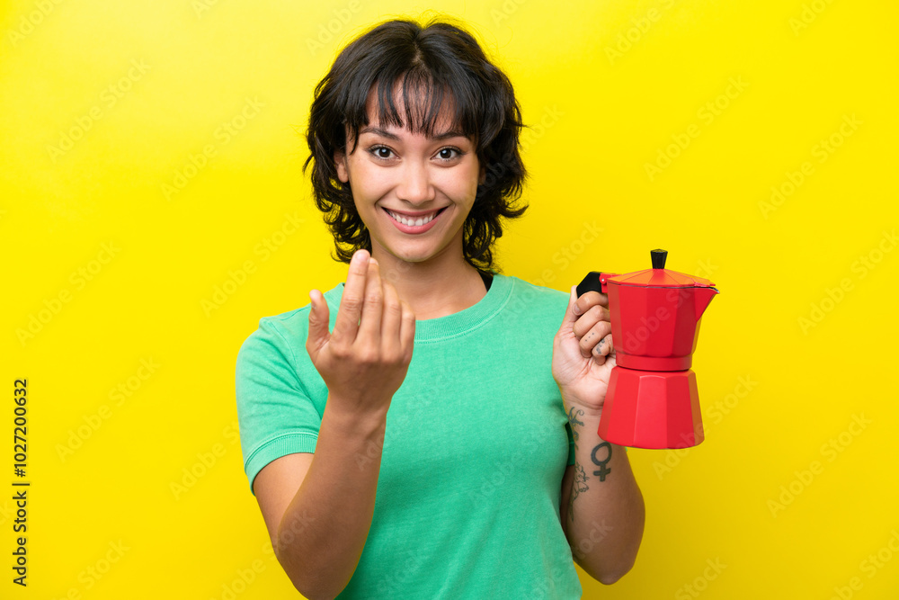 Young Argentinian woman holding coffee pot isolated on yellow background inviting to come with hand. Happy that you came Young Argentinian woman holding coffee pot isolated on yellow background inviting to come with hand. Happy that you came