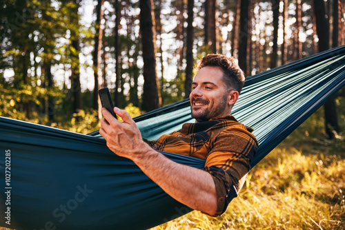 Portrait of carefree man using mobile phone in hammock during afternoon in forest