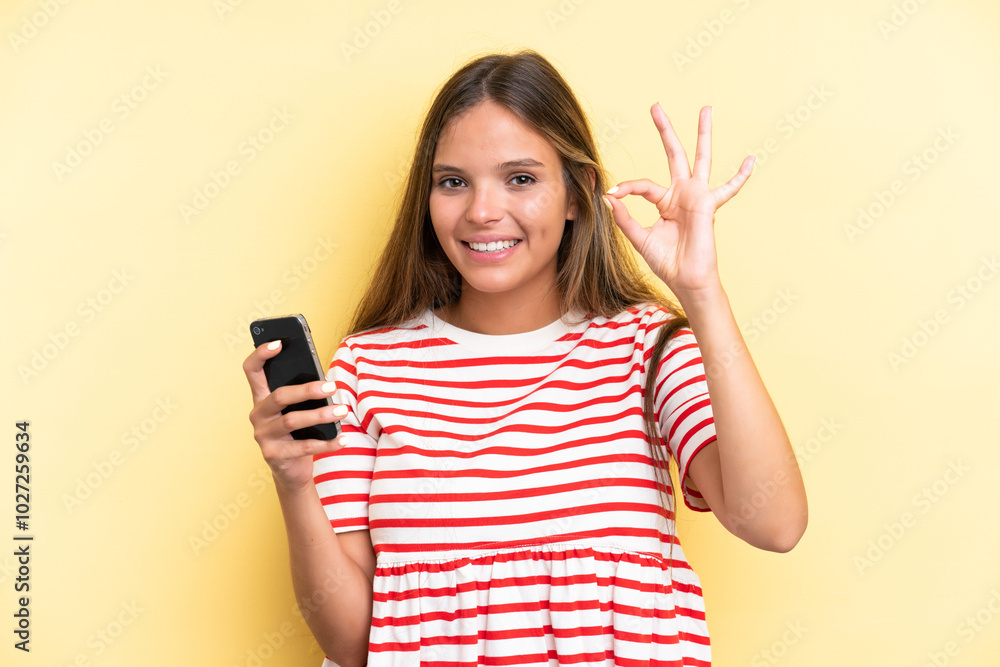 Young caucasian woman isolated on yellow background using mobile phone and doing OK sign Young caucasian woman isolated on yellow background using mobile phone and doing OK sign