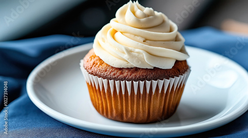 Close-up of a vanilla cupcake with creamy swirled frosting on a white plate, set on a blue fabric surface.