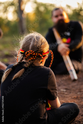 Aboriginal girl listening to man playing didgeridoo in fading light in Australian bushland