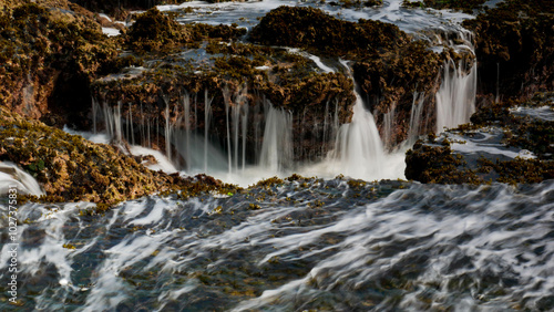 The crashing waves at Karang Bokor Sawarna Beach flowing over the coral rocks become a mini waterfall. The rough texture of the coral rocks and green moss add dimension to the scenery.