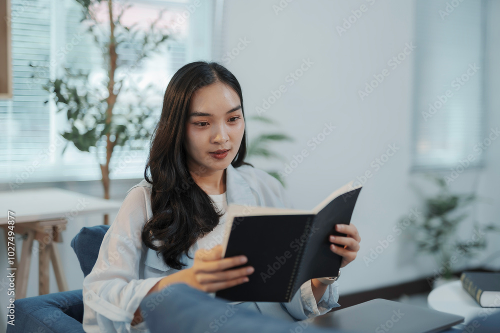 Young asian businesswoman is sitting on a comfortable blue couch in an office lounge, deeply engrossed in reading a book Young asian businesswoman is sitting on a comfortable blue couch in an office lounge, deeply engrossed in reading a book
