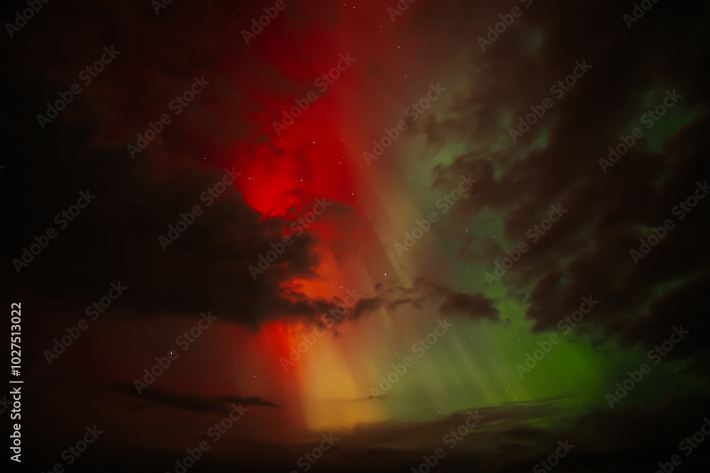 Fototapeta premium A vivid display of the Aurora Borealis lights up the night sky, with striking red, orange, and green hues blending amidst dark clouds. The dramatic contrast creates a surreal and captivating scene.