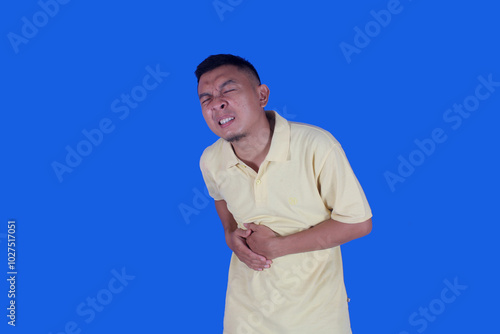 Funny face of asian man wearing t-shirt suffering from stomachache due to diarrhea on blue background
