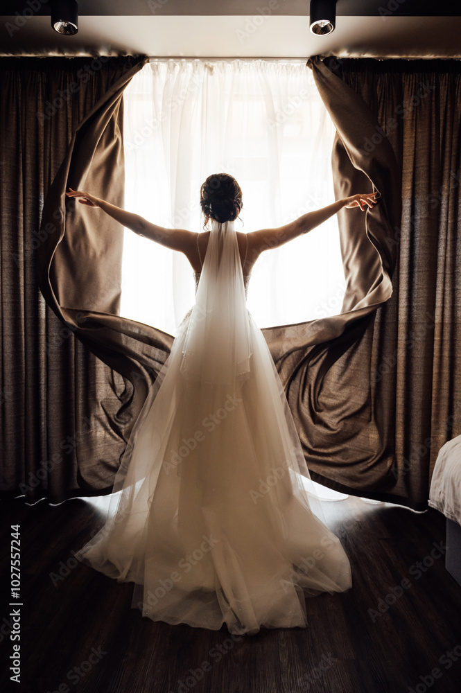 Fototapeta premium The girl stands with her back near the window and opens the curtains with her hands