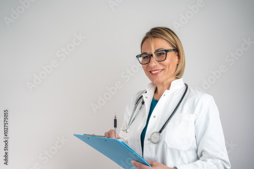 Healthcare consultation. Smiling mature female doctor in medical coat writing in clipboard isolated on white. Mature female doctor on light background. 