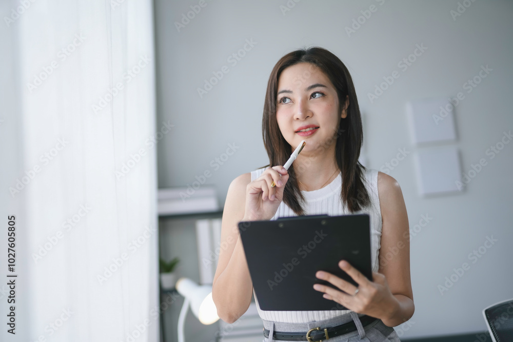 Young businesswoman is holding a clipboard and pen while looking away thoughtfully, generating innovative solutions for her next project Young businesswoman is holding a clipboard and pen while looking away thoughtfully, generating innovative solutions for her next project