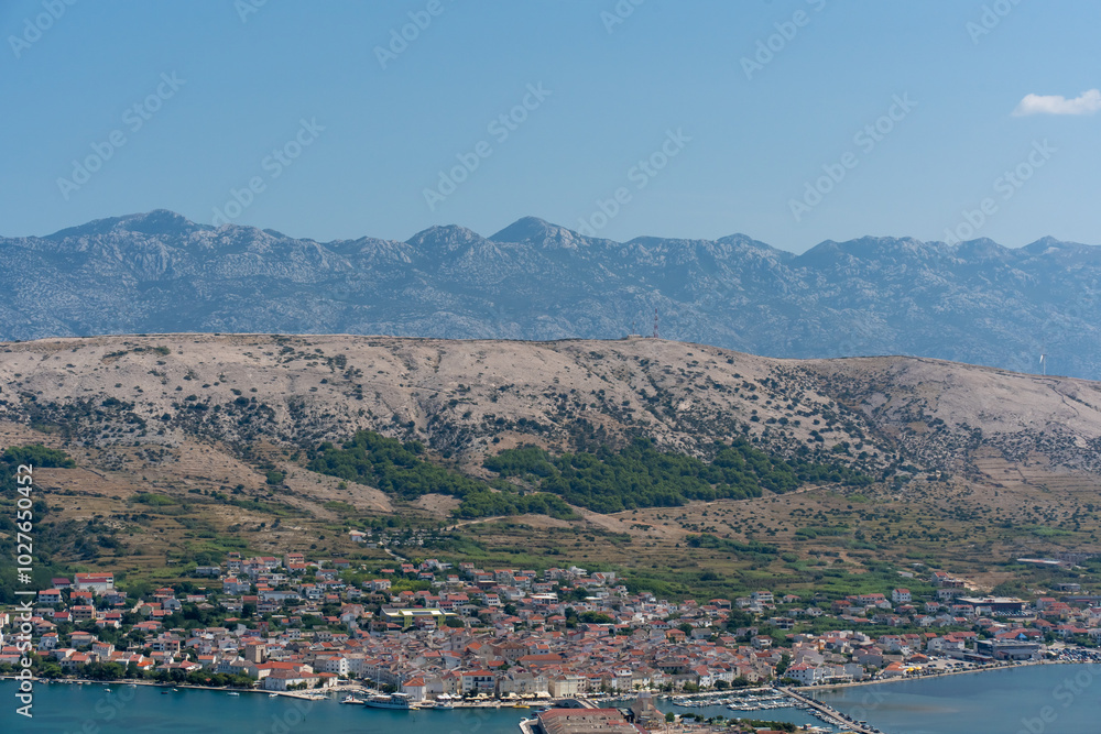 Picturesque coastal town with a mountain range backdrop, showing red-roofed houses, a calm bay, and rugged hills under a clear blue sky. Serene Mediterranean landscape captured from above. Picturesque coastal town with a mountain range backdrop, showing red-roofed houses, a calm bay, and rugged hills under a clear blue sky. Serene Mediterranean landscape captured from above.