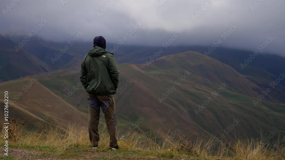 Hiker stands on a mountainside, gazing at a breathtaking mountain vista under a cloudy sky. The scene showcases nature's beauty and the serenity of the wilderness Hiker stands on a mountainside, gazing at a breathtaking mountain vista under a cloudy sky. The scene showcases nature's beauty and the serenity of the wilderness