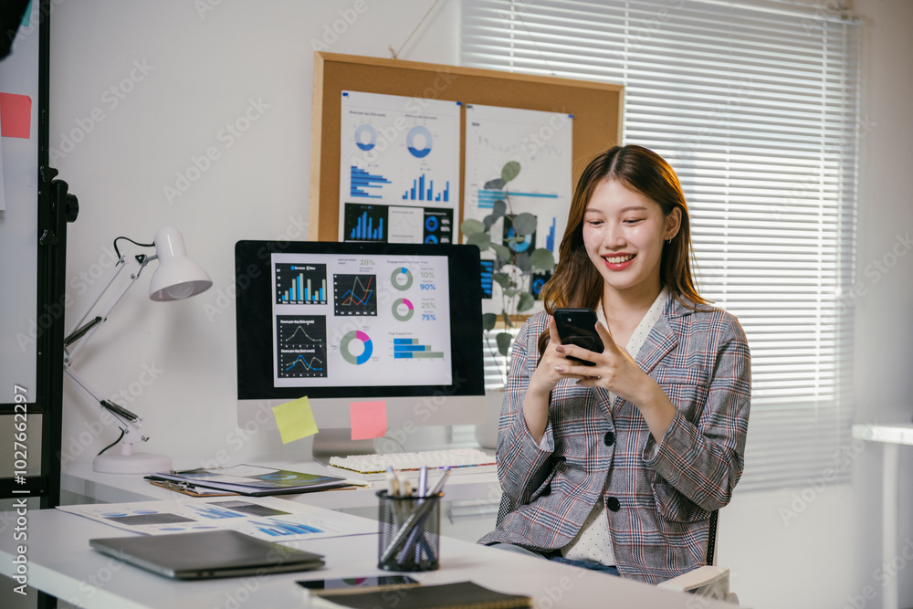 Focused businesswoman in a modern office, surrounded by charts on her computer, takes a break to smile at her smartphone, blending work and relaxation effortlessly Focused businesswoman in a modern office, surrounded by charts on her computer, takes a break to smile at her smartphone, blending work and relaxation effortlessly