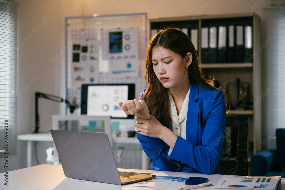 Young manager feeling wrist pain while working on a laptop at her desk in the office. She is massaging her wrist to relieve the pain Young manager feeling wrist pain while working on a laptop at her desk in the office. She is massaging her wrist to relieve the pain