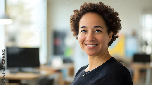 A smiling woman with curly hair poses in a modern office environment.