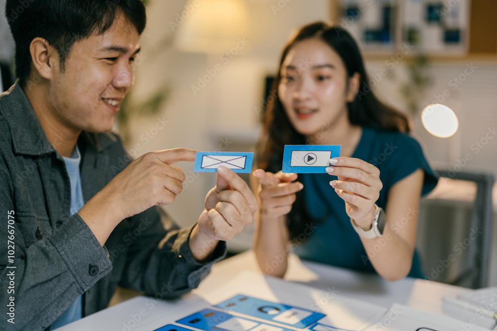 Two ui/ux designers smiling and discussing about new features for an app, holding paper prototypes of the user interface Two ui/ux designers smiling and discussing about new features for an app, holding paper prototypes of the user interface