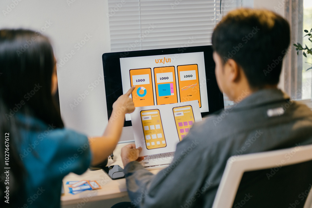 Two young ui/ux designers are working together in the office, discussing over design prototypes on paper and computer screen Two young ui/ux designers are working together in the office, discussing over design prototypes on paper and computer screen