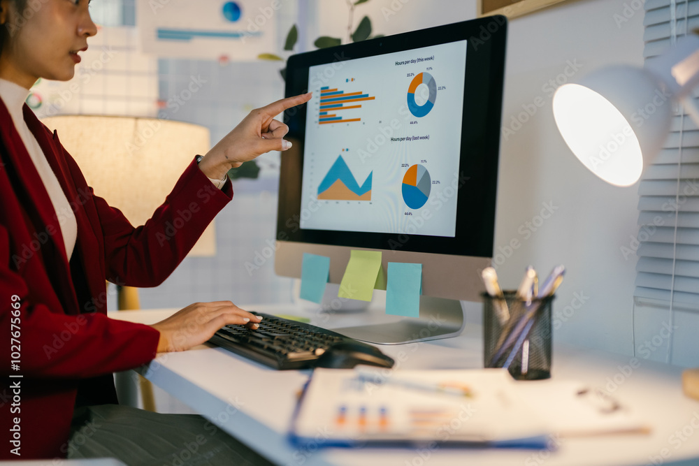 Fototapeta premium Businesswoman analyzing marketing data on a computer screen, pointing at charts and graphs. She is working late at night in a modern office
