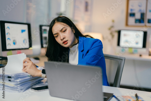 Stressed businesswoman overwhelmed by paperwork in modern office, facing deadlines and busy day, analyzing charts and graphs on laptop