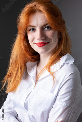 Portrait of a red-haired girl in a white shirt. Woman smiling in studio against gray background.