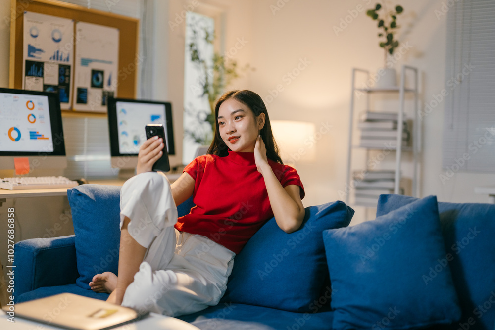 Young asian woman sitting comfortably on a blue couch in her modern home office. Taking a break and using her smartphone for a video call. Enjoying the evening in her cozy apartment Young asian woman sitting comfortably on a blue couch in her modern home office. Taking a break and using her smartphone for a video call. Enjoying the evening in her cozy apartment